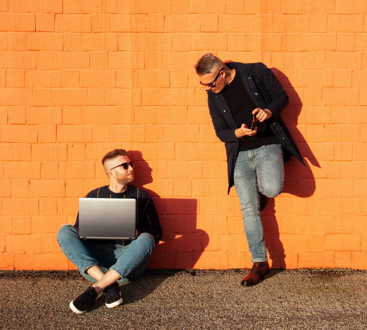 One man sitting down with a laptop and one man standing with a cell phone both in front of an orange wall.
